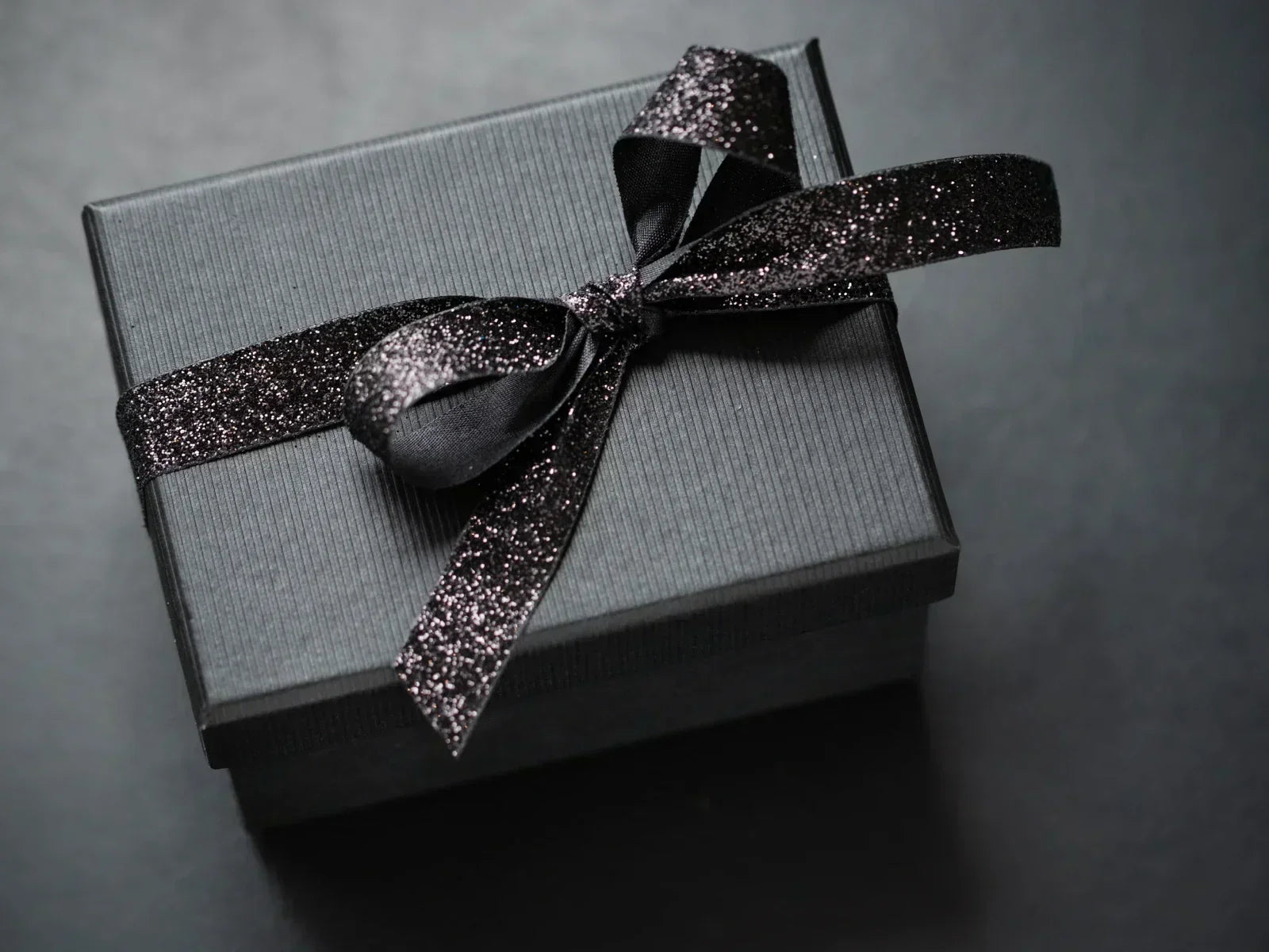 A dark luxury gift box with a glittering black ribbon on a dark background — representing the moment before opening when elegant packaging already communicates care and intention